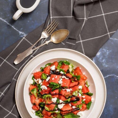 Plate of watermelon salad with feta, mint, and balsamic glaze. A fork and spoon rest beside on a checkered cloth, and a small jug of balsamic dressing is placed nearby.