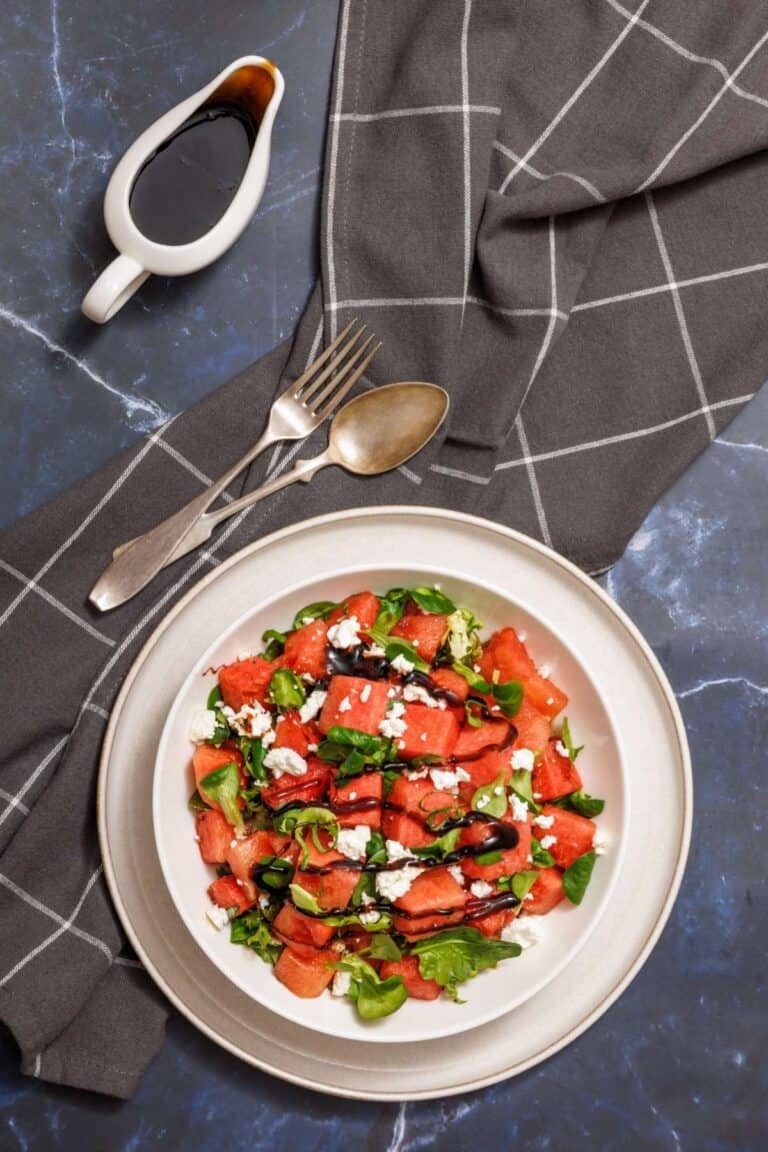 Plate of watermelon salad with feta, mint, and balsamic glaze. A fork and spoon rest beside on a checkered cloth, and a small jug of balsamic dressing is placed nearby.