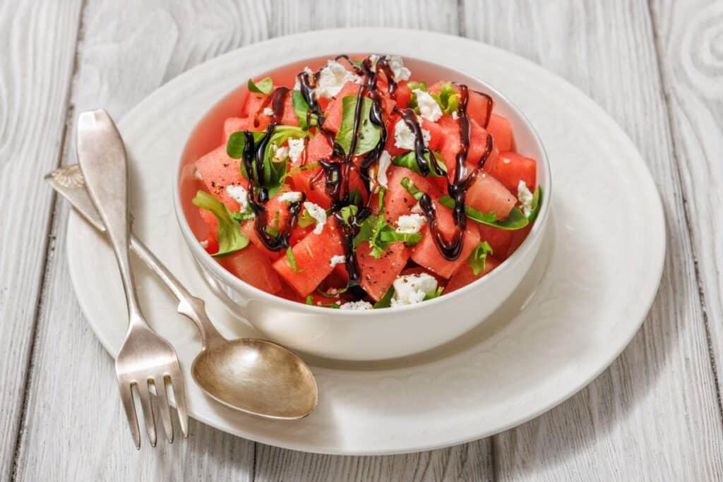Bowl of watermelon salad with feta, greens, and balsamic glaze on a white plate. Fork and spoon beside the bowl on a wooden surface.