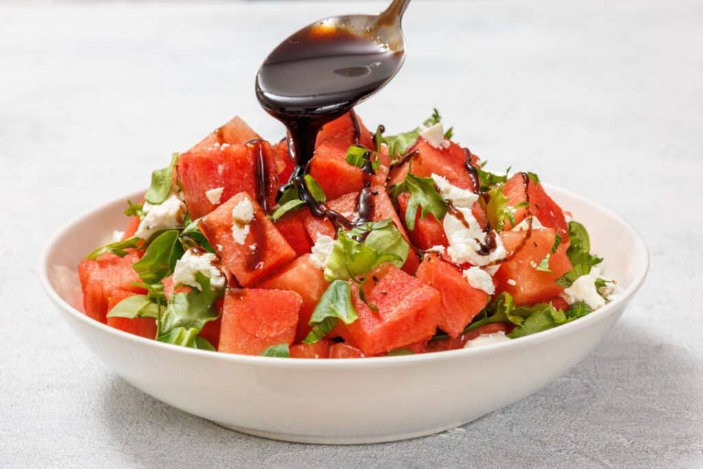 A spoon drizzles balsamic glaze over a salad with watermelon cubes, crumbled feta, and arugula in a white bowl on a light surface.