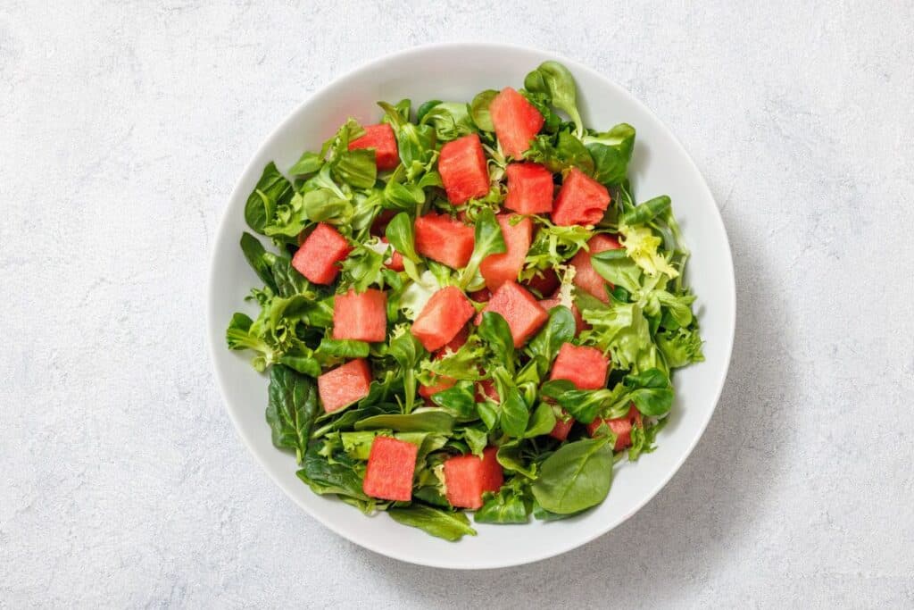 A white bowl contains a salad with mixed greens and diced watermelon on a light gray textured background.