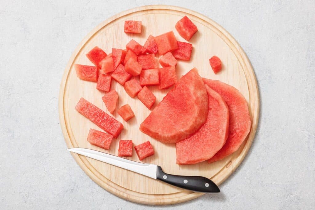 Chopped watermelon and slices on a round wooden cutting board with a knife.