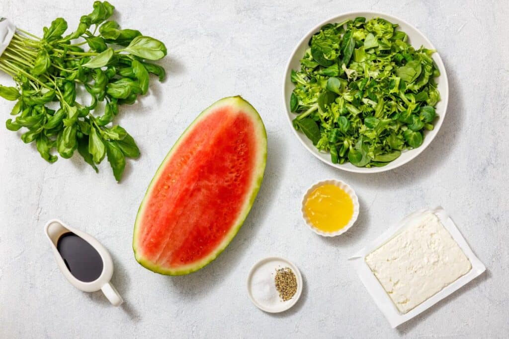 Ingredients for a salad: half a watermelon, arugula in a bowl, feta cheese, olive oil, vinegar, black pepper, and fresh greens on a light surface.