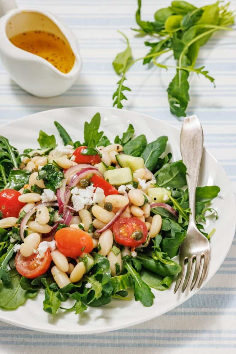 A fresh White Bean Salad with arugula, cherry tomatoes, white beans, cucumber, red onion, feta cheese, and capers on a striped cloth. A fork rests on the plate; dressing is in a small jug nearby.