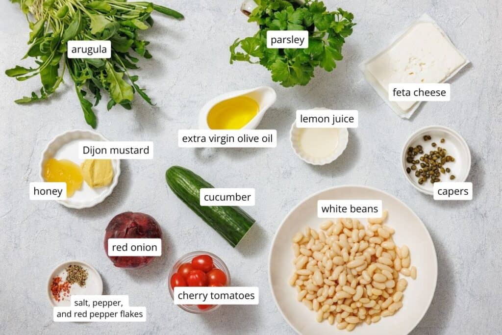 Ingredients for White Bean Salad are arranged on a surface: arugula, parsley, feta cheese, cucumber, red onion, cherry tomatoes, white beans, capers, Dijon mustard, honey, lemon juice, olive oil, spices.
