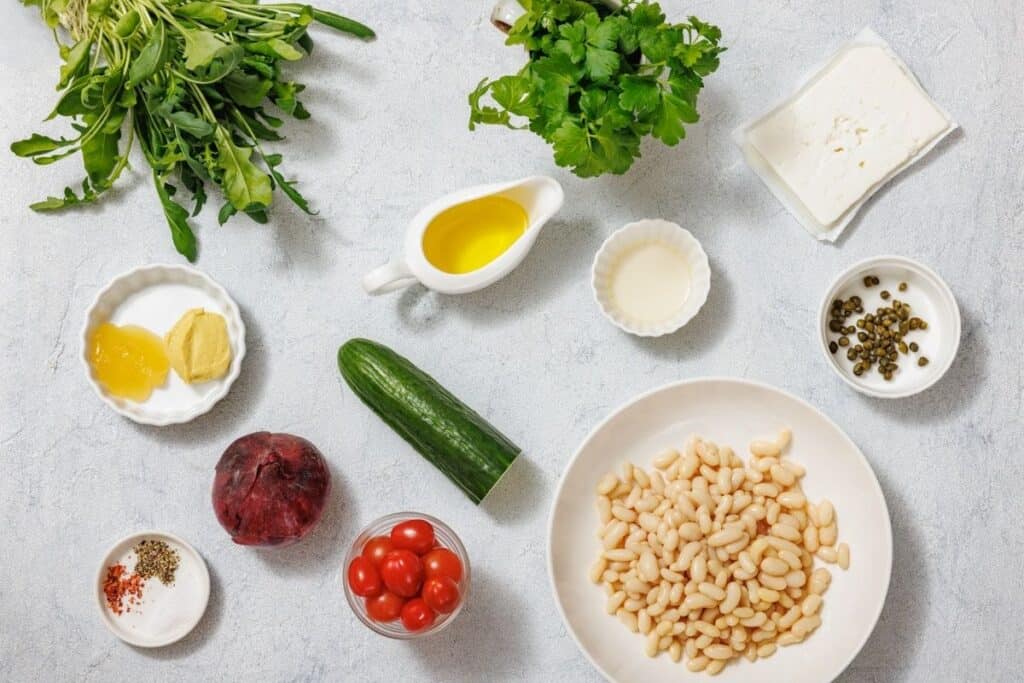 Ingredients for White Bean Salad on a surface include arugula, parsley, feta, olive oil, vinegar, capers, cucumber, beet, cherry tomatoes, white beans, seasonings, honey, and mustard.