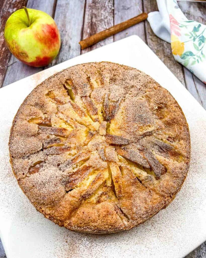 Round apple cake with cinnamon sugar topping on a white plate. An apple and a cinnamon stick are beside it on a wooden surface.