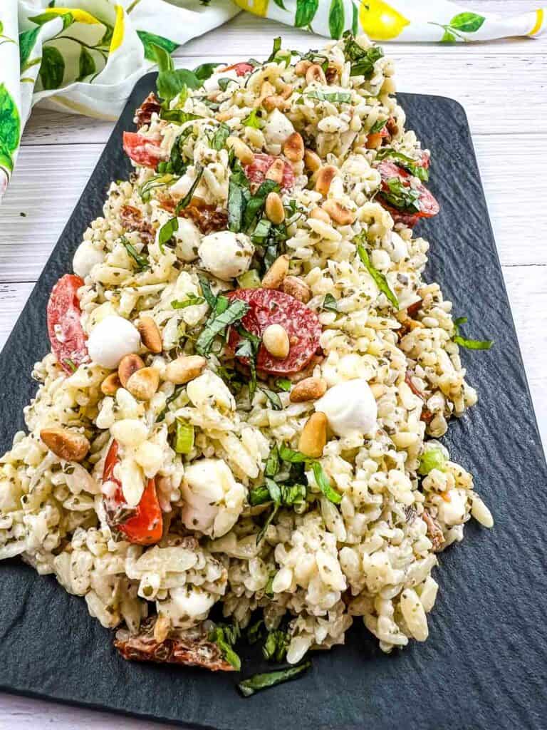 A platter of rice salad with tomatoes, mozzarella balls, pine nuts, and herbs, served on a rectangular black plate.