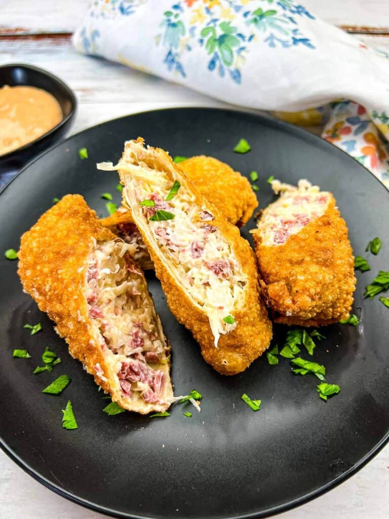 Three crispy egg rolls filled with a creamy mixture cut open on a black plate, garnished with green herbs, with a sauce dish in the background.