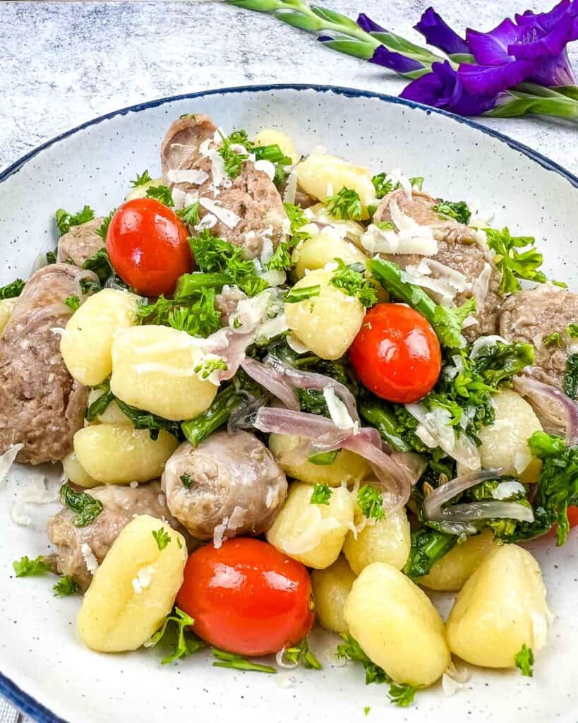 A white bowl filled with gnocchi, cherry tomatoes, meat, onions, and greens, topped with grated cheese. A purple flower is visible in the background.