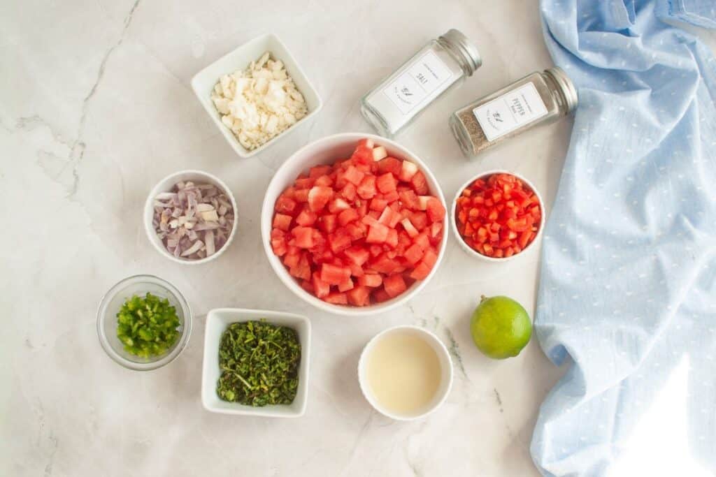 Overhead shot of ingredients for watermelon salad laid out in small bowls on a white marble surface, including diced watermelon, red bell pepper, shallots, feta cheese, jalapeno, cilantro, lime juice, lime, salt, and pepper. A blue cloth is draped on the side.