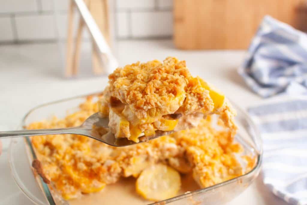 A close-up of a spoonful of yellow squash casserole held over a glass baking dish filled with more casserole, on a kitchen counter.