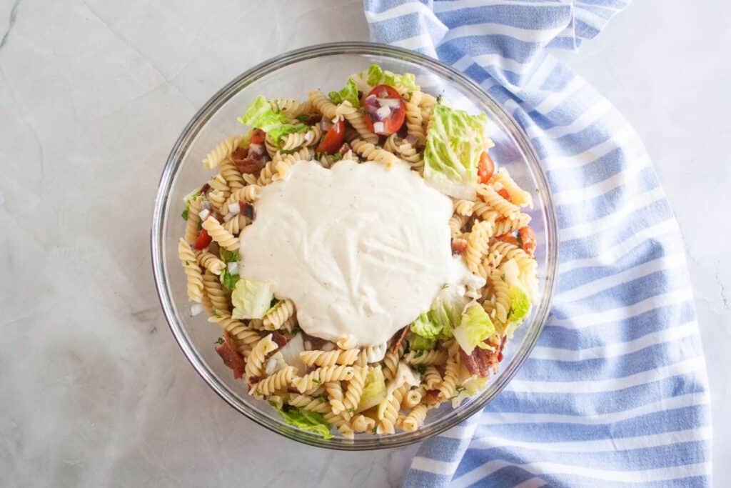 Overhead shot of a glass bowl filled with pasta salad, featuring spiral pasta, tomatoes, bacon, lettuce, and red onion, with a dollop of creamy dressing on top. A blue striped cloth is visible on the side.