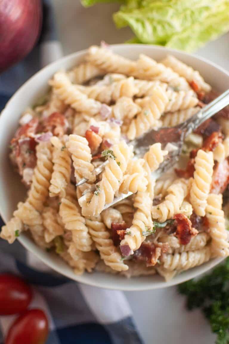 A fork lifts a portion of BLT pasta salad with spiral pasta, creamy dressing, and pieces of bacon from a white bowl. Red onions and herbs are also visible in the salad. A blue checkered cloth and cherry tomatoes are in the background.
