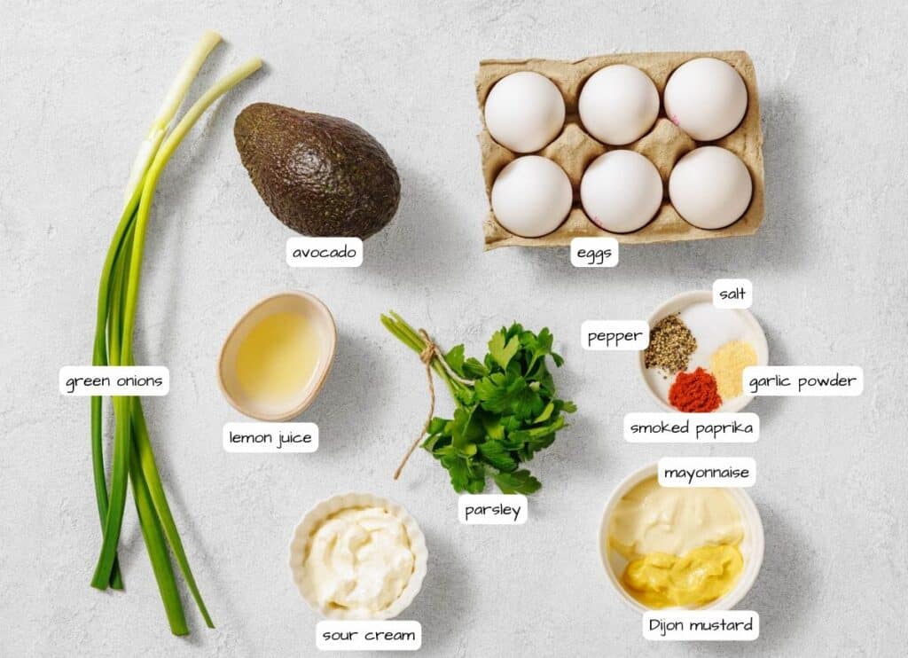 Overhead shot of avocado egg salad ingredients, including eggs, avocado, herbs, mayonnaise, mustard, and spices, arranged on a light surface with labels for each ingredient.