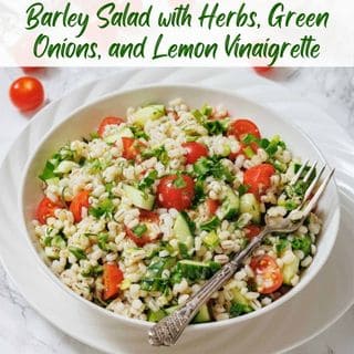 A bowl of barley salad with cherry tomatoes, green onions, cucumber, and herbs, served with a fork on a white plate.