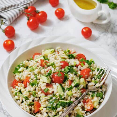 A bowl of barley salad with cherry tomatoes, cucumber, herbs, and a fork, set on a white plate with a jug of dressing and fresh tomatoes in the background.