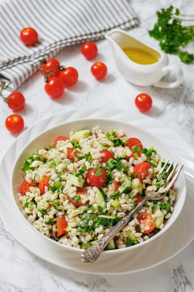 A bowl of barley salad with cherry tomatoes, cucumber, herbs, and a fork, set on a white plate with a jug of dressing and fresh tomatoes in the background.