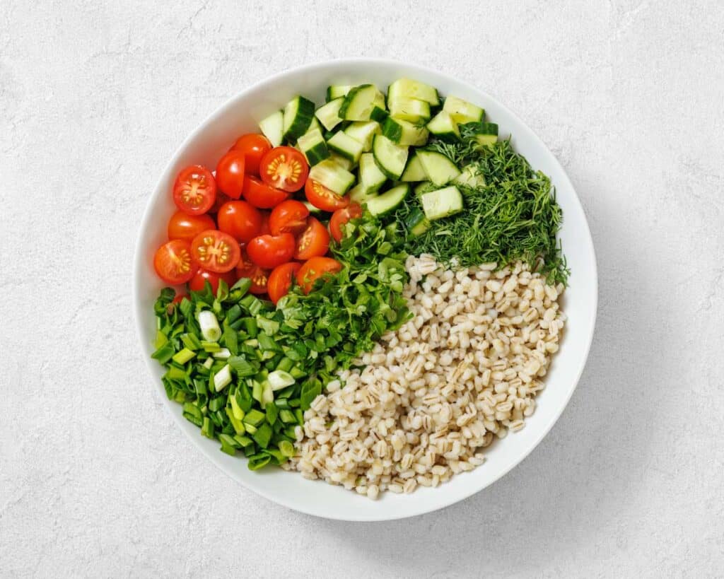 A white bowl containing sections of cherry tomatoes, chopped cucumber, dill, green onions, parsley, and cooked barley on a light background.