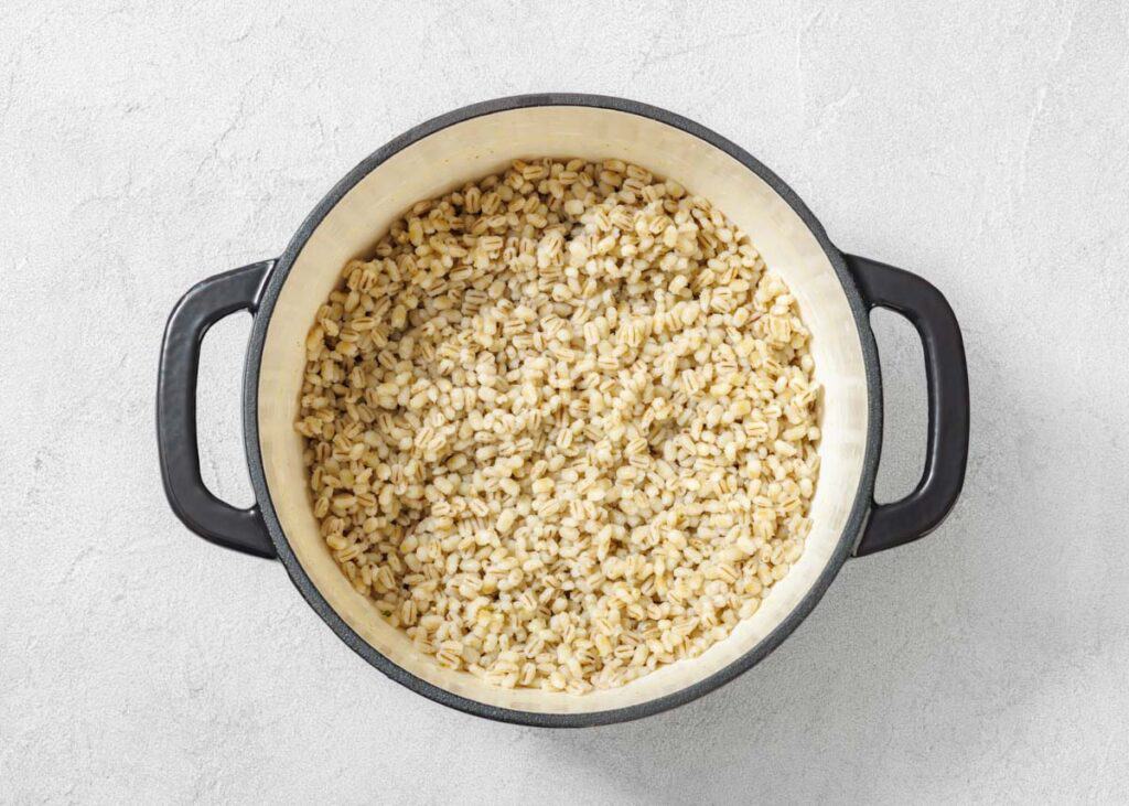 A black-handled pot on a light surface filled with cooked barley grains.