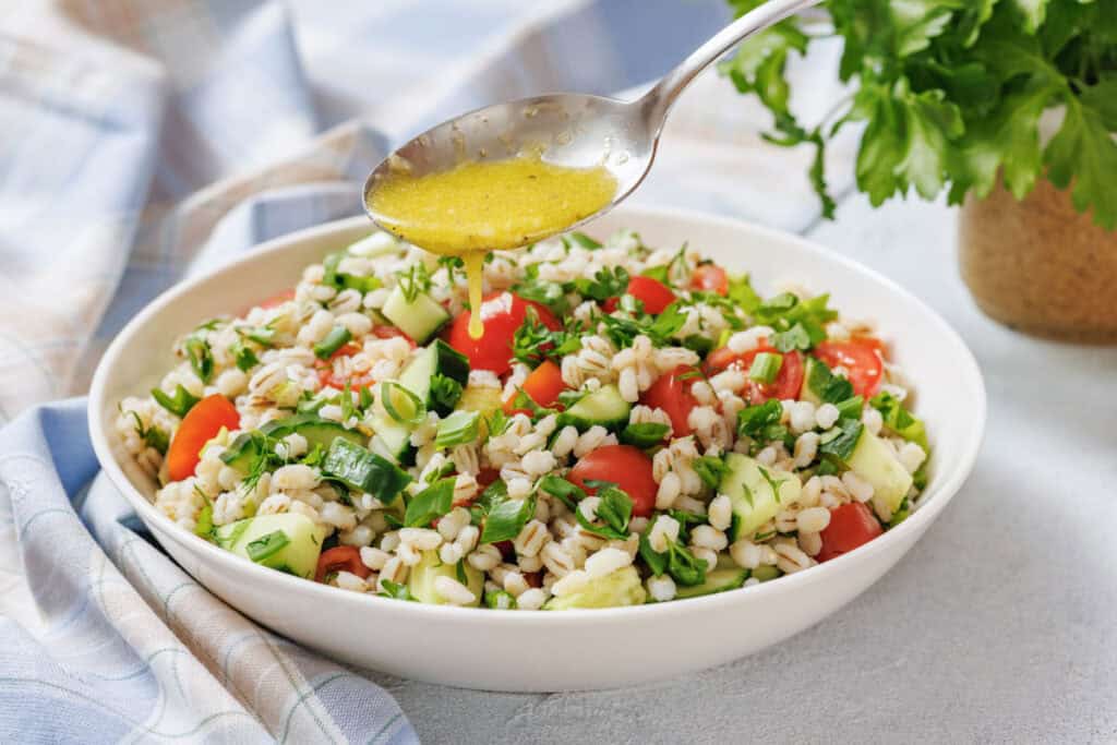 A spoon pours yellow dressing over a bowl of barley salad with tomatoes, cucumbers, green onions, and herbs on a table with a blue-checked cloth.