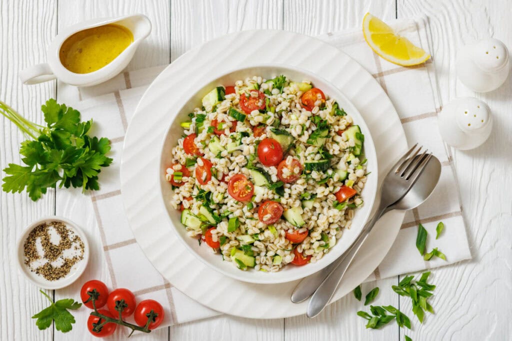 A bowl of barley salad with cherry tomatoes, cucumber, parsley, and green onions, surrounded by dressing, lemon wedge, parsley, seasonings, and utensils on a white tabletop.
