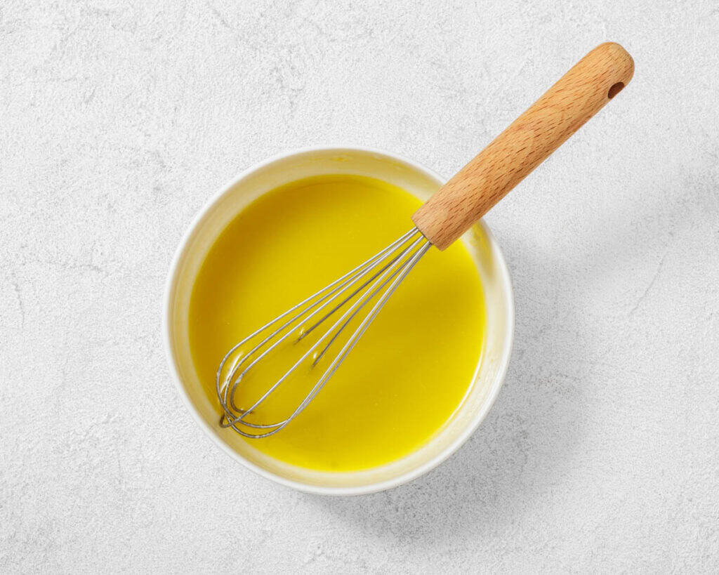 A whisk rests in a bowl filled with yellow liquid mixture on a light textured surface.