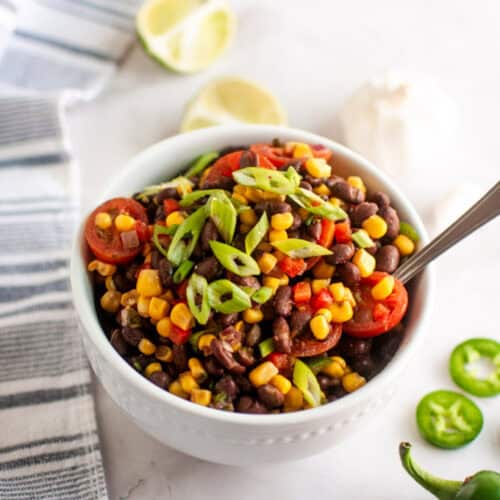 A white bowl filled with a salad of black beans, corn, cherry tomatoes, and sliced green onions, with a spoon inside; lime halves and sliced jalapeños are nearby.