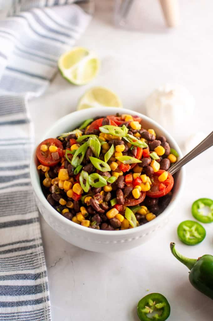 A white bowl filled with a salad of black beans, corn, cherry tomatoes, and sliced green onions, with a spoon inside; lime halves and sliced jalapeños are nearby.