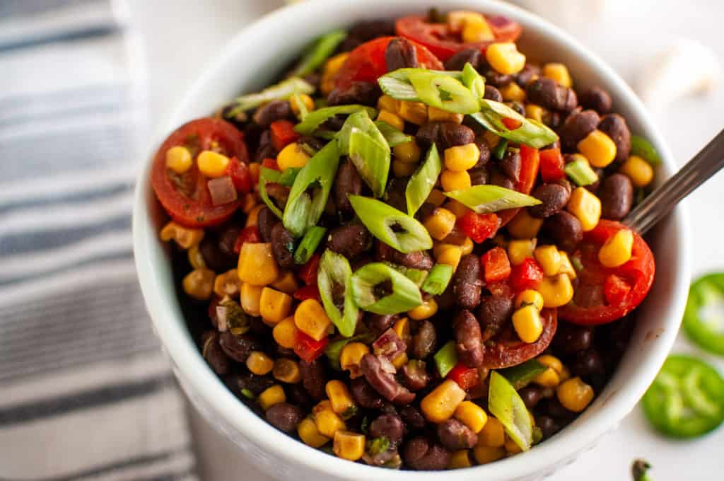 A bowl of salad with black beans, corn, cherry tomatoes, red bell pepper, and sliced green onions, with a spoon inside.