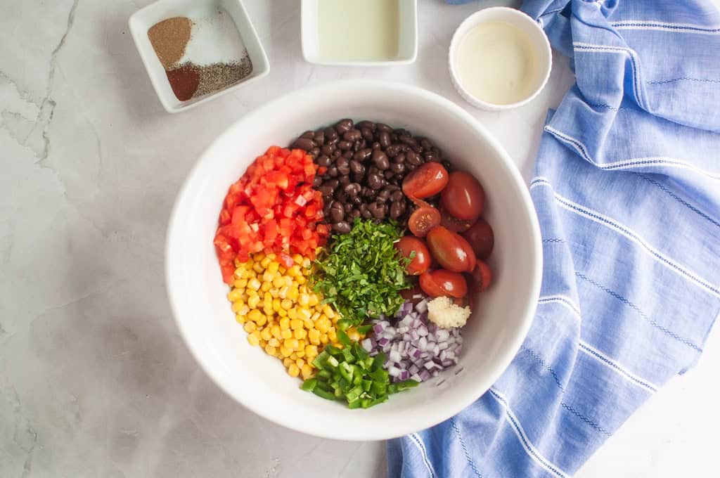 A white bowl contains black beans, diced tomatoes, sliced cherry tomatoes, corn, chopped cilantro, jalapeño, red onion, and minced garlic. Small bowls with spices and dressing are nearby.