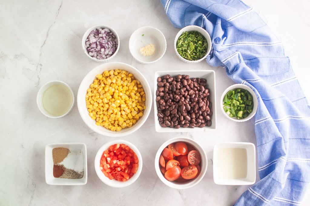 Various ingredients in bowls on a white surface, including black beans, corn, chopped vegetables, spices, and a blue striped cloth.