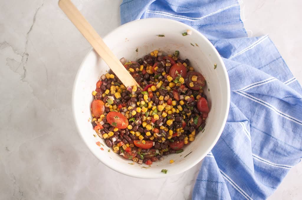 A white bowl filled with a black bean, corn, and tomato salad, with a wooden spoon inside, sits next to a blue and white striped cloth on a light surface.