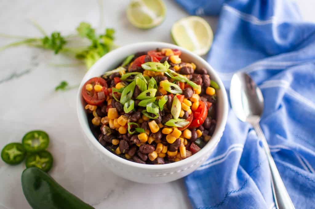 A bowl of black bean and corn salad with sliced tomatoes and green onions, surrounded by fresh cilantro, lime halves, jalapeño, and a blue cloth napkin with a spoon.