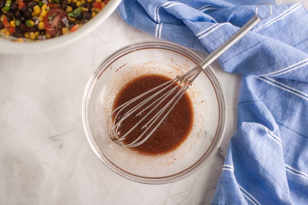 A glass bowl with brown vinaigrette and a whisk sits on a marble surface next to a blue cloth and a partially visible bowl of salad.