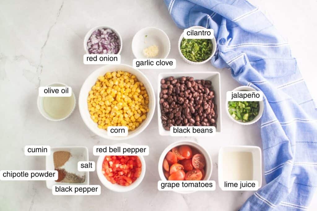Assorted ingredients in bowls for a salsa, including corn, black beans, grape tomatoes, red onion, cilantro, jalapeño, red bell pepper, spices, olive oil, and lime juice, on a white surface.