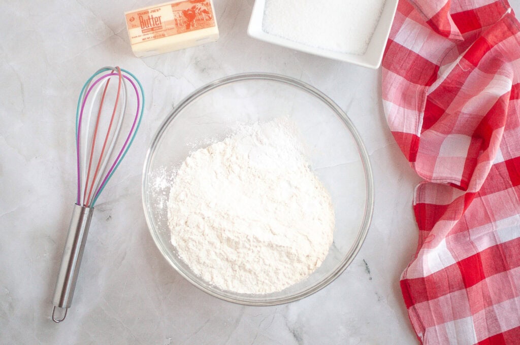 A glass bowl with flour, a stick of butter, a whisk, a square dish of sugar, and a red-checkered cloth on a light countertop.