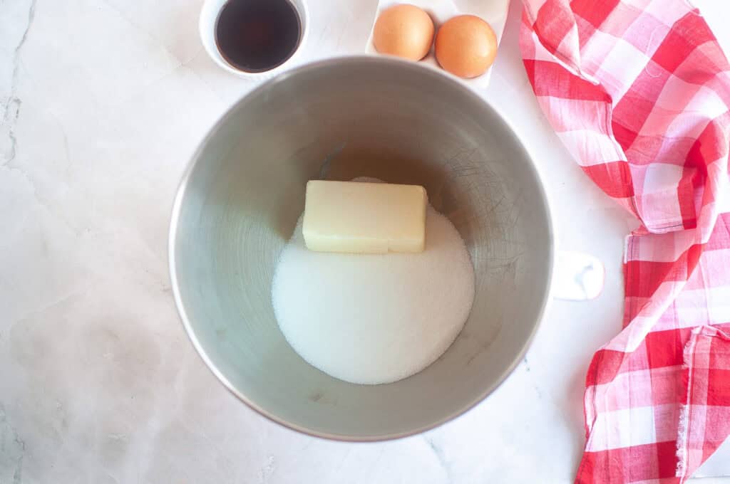A mixing bowl with a stick of butter and granulated sugar inside, next to two eggs, a small bowl of vanilla, and a red and white checkered cloth on a marble surface.