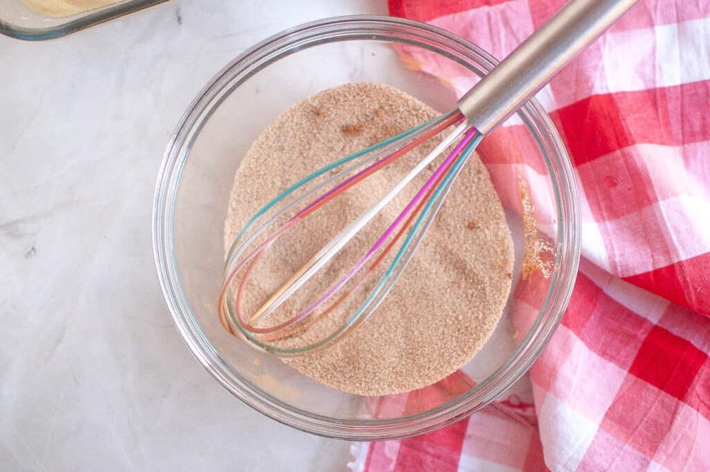 A glass bowl filled with cinnamon sugar and a metal whisk, next to a red and white checkered cloth on a marble surface.
