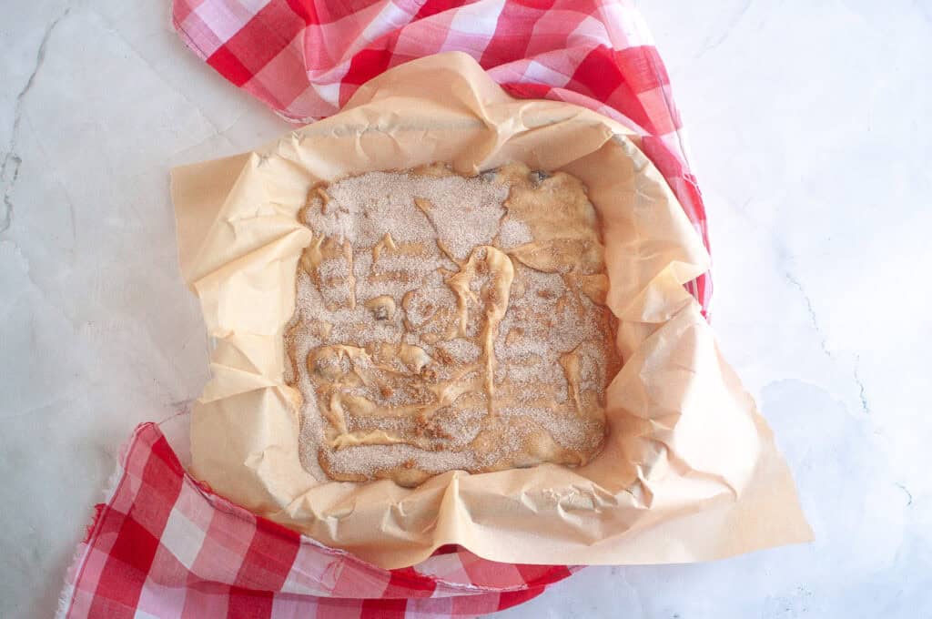 Unbaked dough spread in a parchment-lined baking dish, topped with sugar, placed on a red and white checkered cloth.