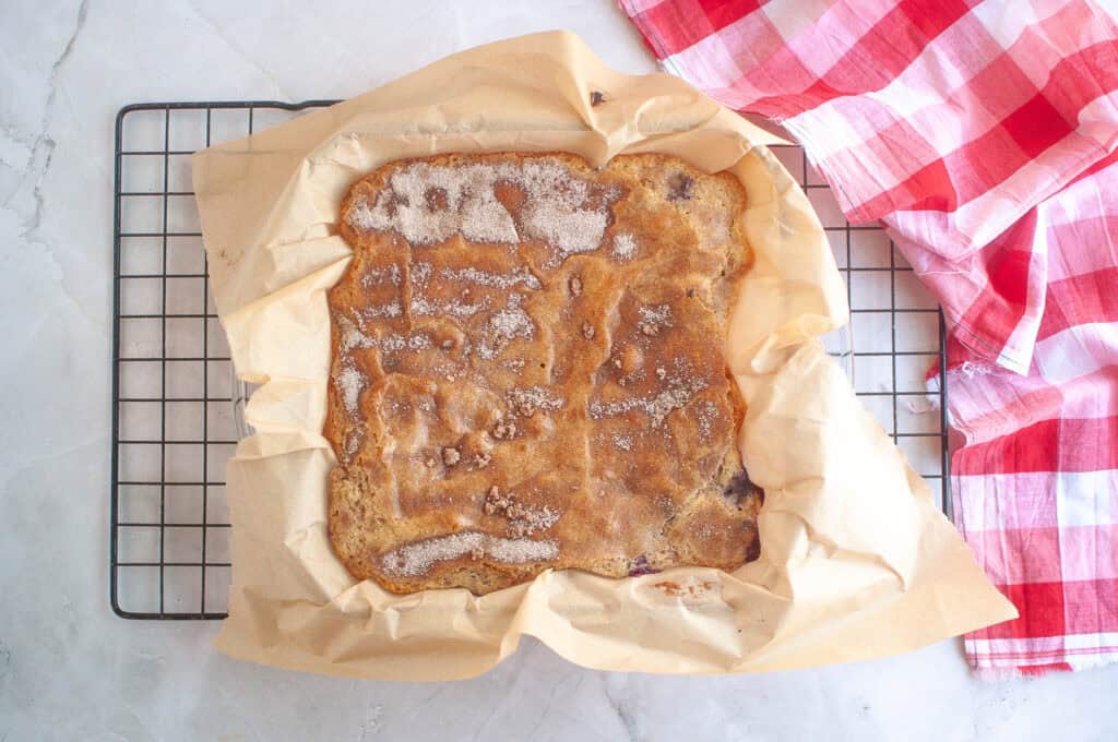 A baked square dessert in parchment paper sits on a cooling rack beside a red and white checkered cloth.
