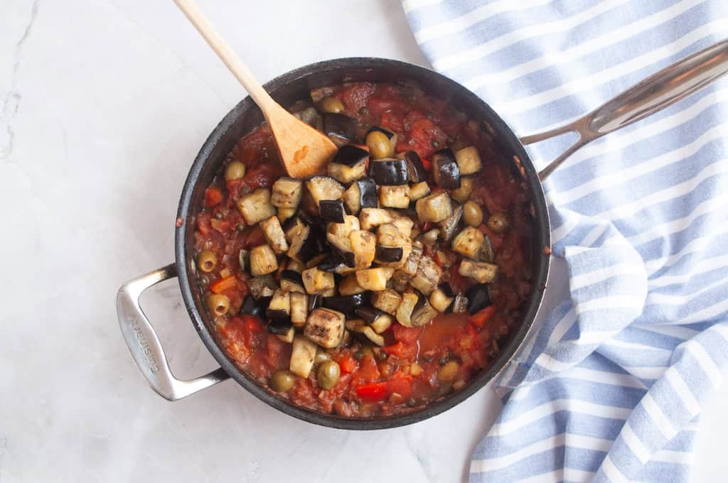A pan filled with tomato-based stew, diced eggplant, green olives, and red peppers, with a wooden spoon resting inside; a striped cloth lies nearby.