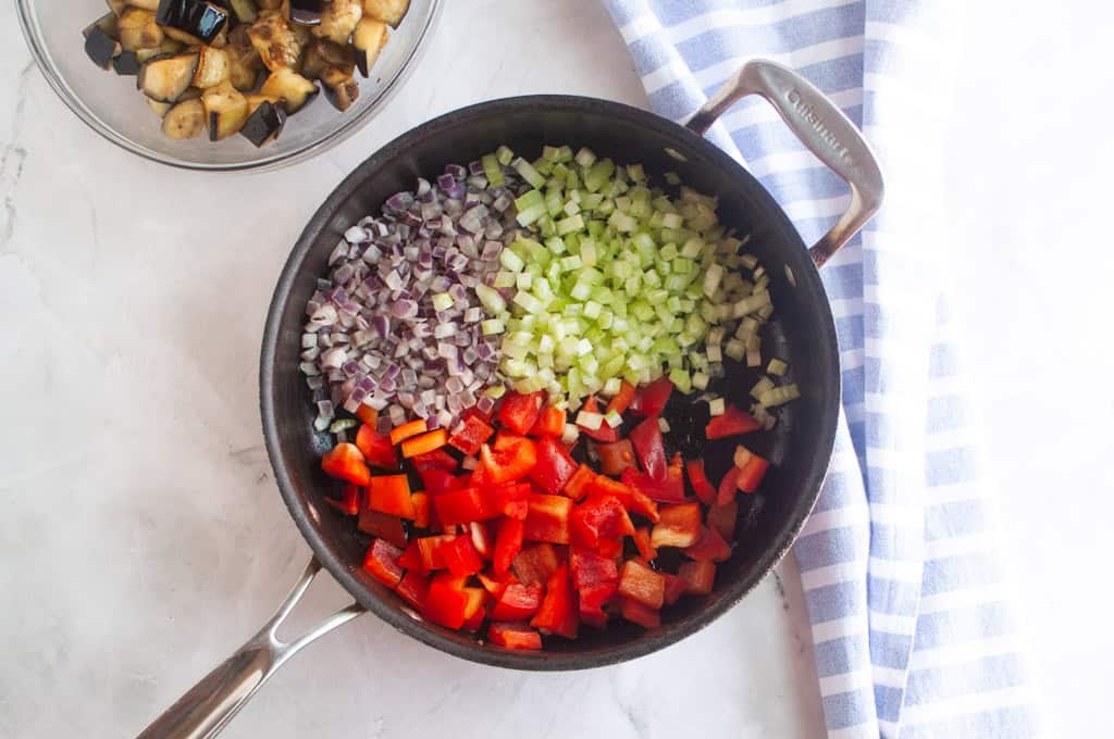 A skillet with diced red bell pepper, celery, and red onion, with a bowl of cooked eggplant and a striped towel beside it on a marble surface.