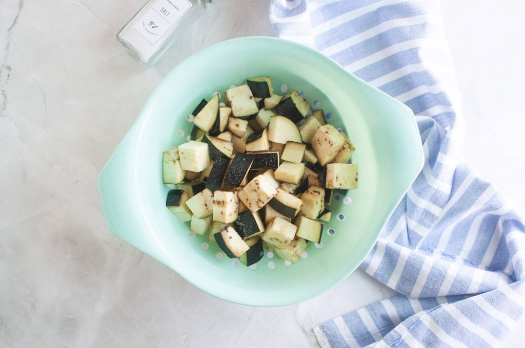 Cubed eggplant pieces in a light green colander, next to a salt container and a blue and white striped dish towel on a light surface.