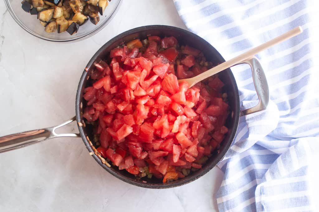 A saucepan filled with diced tomatoes and a wooden spoon sits on a countertop next to a blue and white striped towel and a bowl of roasted vegetables.