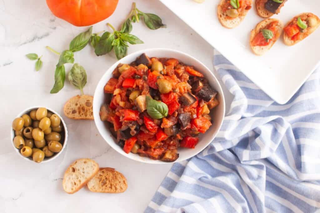 A bowl of ratatouille surrounded by green olives, basil, sliced bread, and bruschetta on a plate, set on a white surface with a striped cloth.