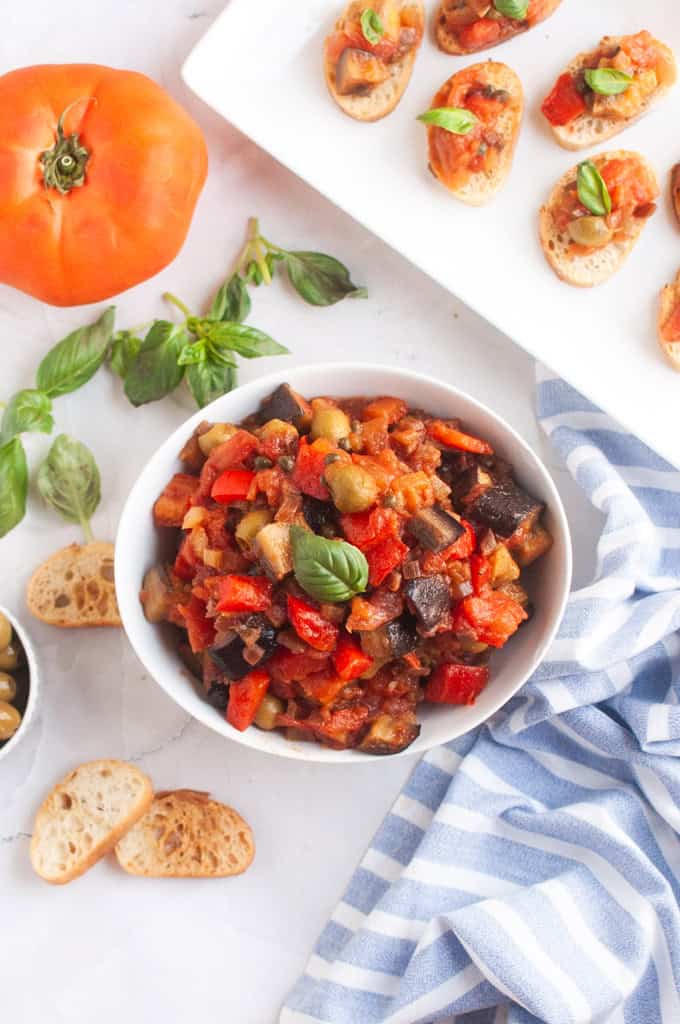 A bowl of Caponata garnished with basil is on a table next to a striped cloth, sliced baguette, a tomato, and fresh basil.