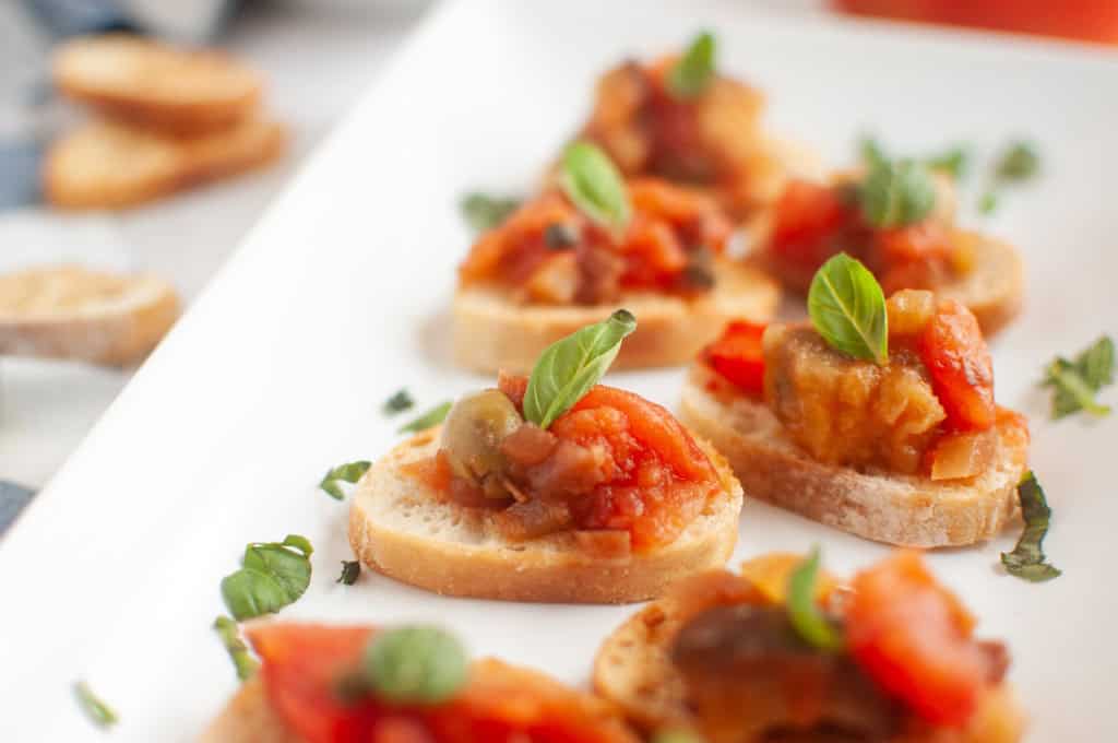 Slices of baguette topped with a tomato-based vegetable mixture and garnished with fresh basil leaves, arranged on a white platter.