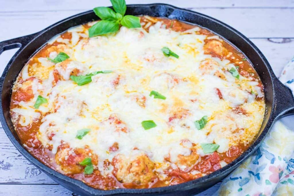 A cast iron skillet filled with meatballs in tomato sauce, topped with melted cheese and garnished with fresh basil leaves.