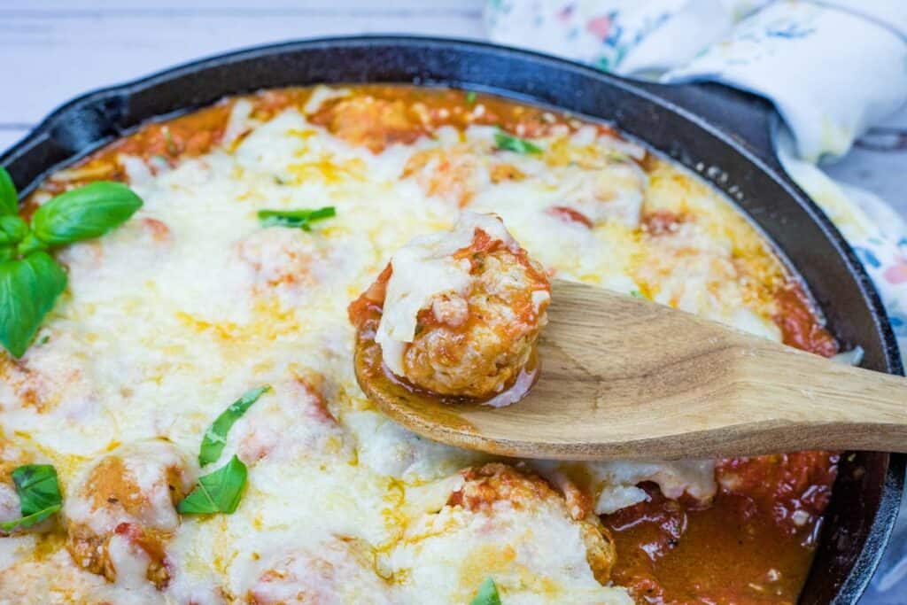 A wooden spoon holds a cheesy baked meatball above a cast iron skillet filled with meatballs in tomato sauce and melted cheese, garnished with fresh basil leaves.